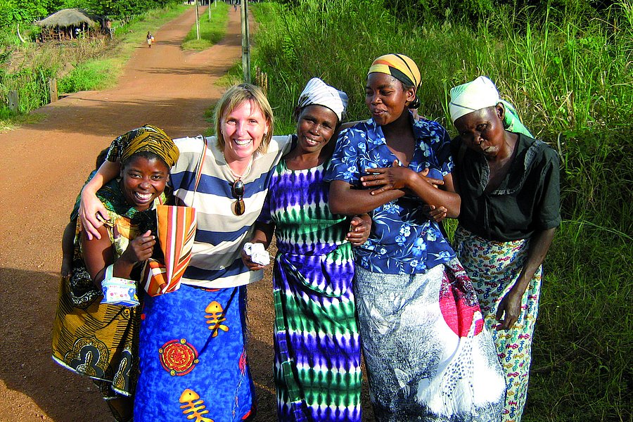 Eine Frau in gestreiftem Shirt posiert lächelnd mit vier einheimischen Frauen in farbenfrohen traditionellen Kleidern auf einem ländlichen Weg.