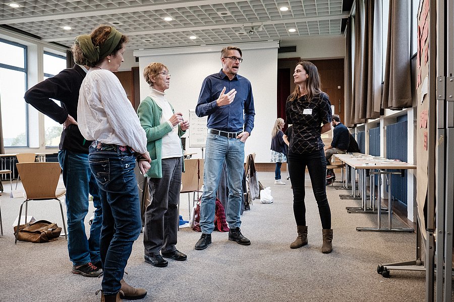 Eine Gruppe von fünf Personen diskutiert in einem Seminarraum. Im Hintergrund sind Stellwände und ein Beamer zu sehen.
